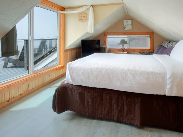 A cozy attic bedroom with a big bed, white linens, a brown bed skirt, sloped ceiling, wood accents, a desk and TV, and a large window overlooking a deck.