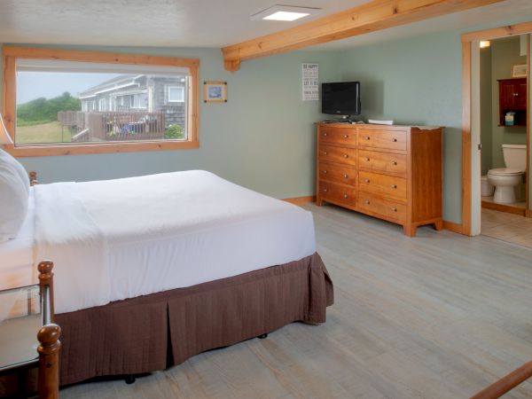 A cozy hotel room with a large bed, light wood floors, a dresser with a TV, a window view of a cottage-like exterior, and an open ensuite bathroom.