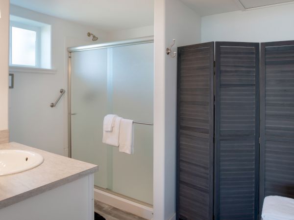 A bright bathroom with a glass shower enclosure, white sink counter, towel on a rail, small window, and a dark folding room screen on the right.