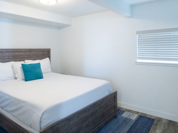 A tidy bedroom with a wooden bed, white bedding, a teal accent pillow, a window with blinds, and a blue-toned rug.