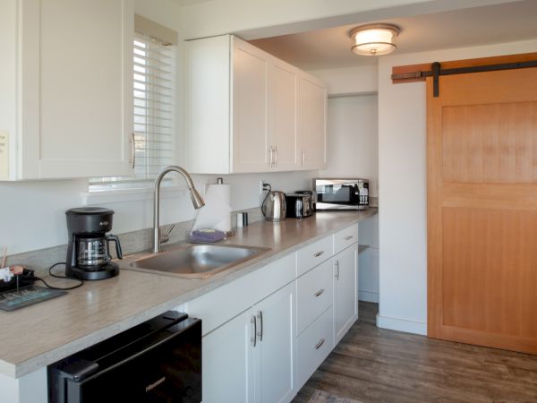 A compact kitchen with white cabinets, a stainless sink, coffee maker, toaster, and various small appliances on a light countertop; a wooden sliding barn door at the end.