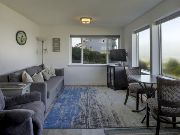 A bright living room with a gray sofa, blue area rug, wall clock, and a small dining table by large windows; a TV on a stand and a view outside.