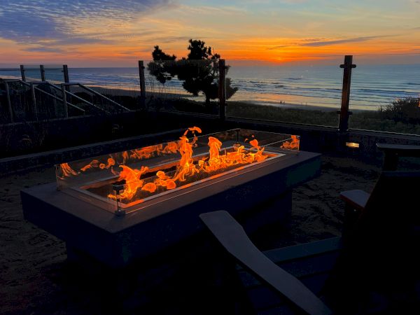 A lit fire pit on a terrace near the coast at sunset, with a tree and fence in the background along the shore.