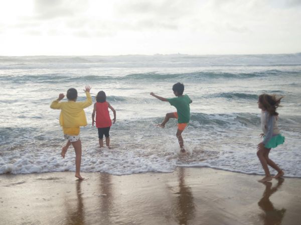 Four kids play at the beach, running near the shoreline as gentle waves roll in under a bright, breezy sky.