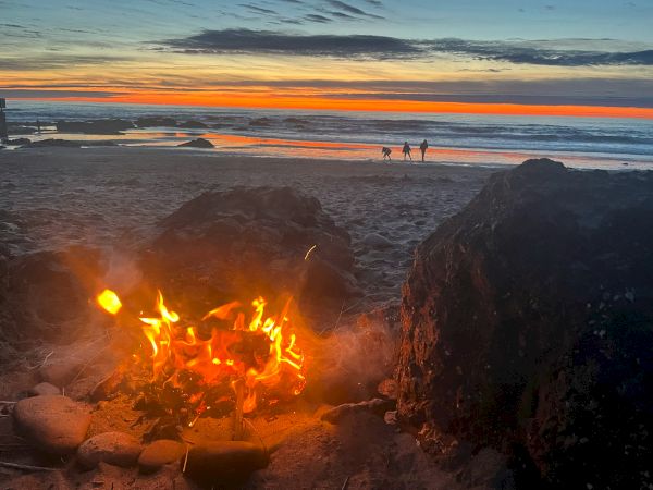 A beach bonfire glows beside rocks at sunset, silhouettes of people near the water as the sky fades to orange and blue.