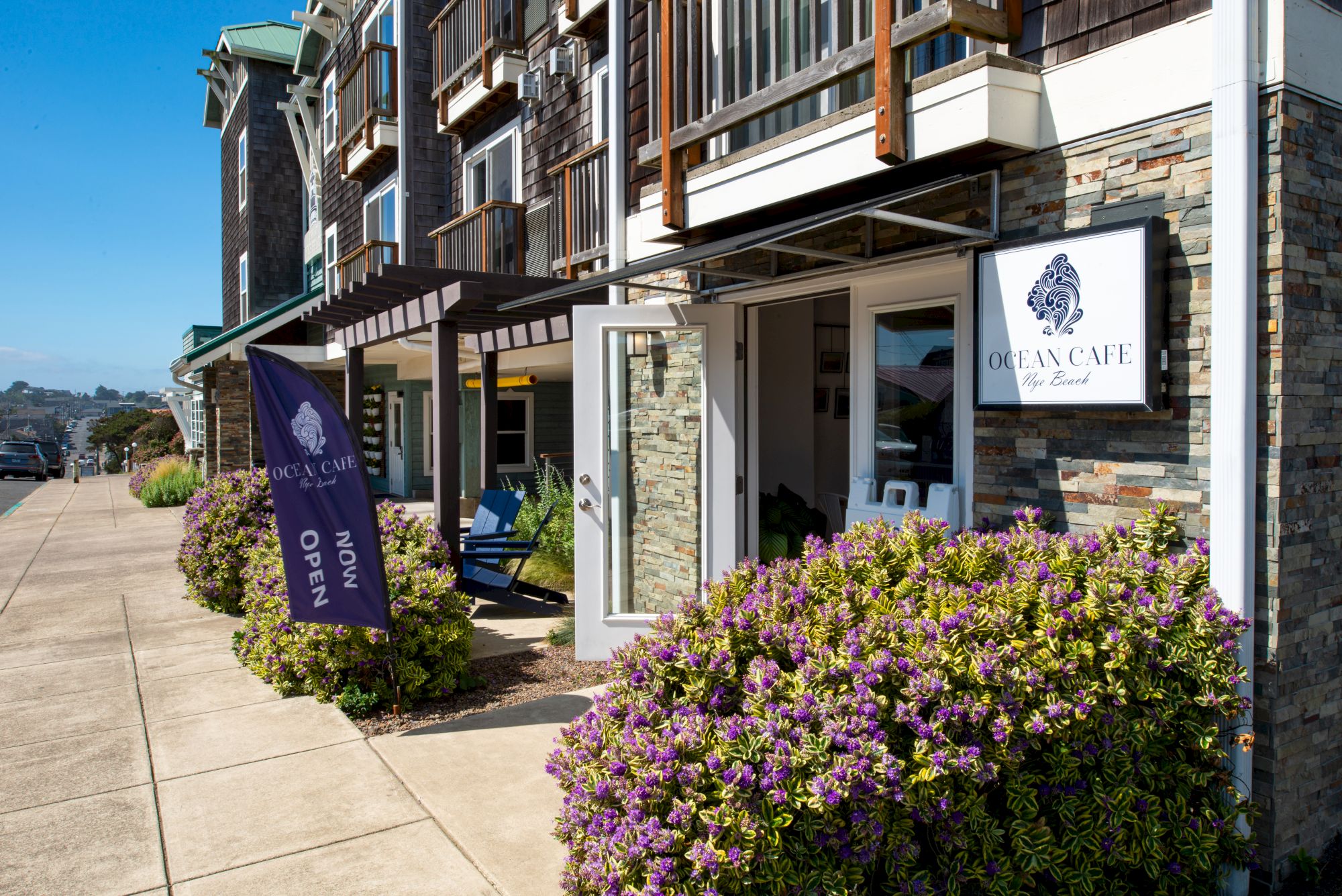 A street view of a modern building with stone siding, hanging awnings, a sign for "GlenCaine" or similar, purple flowering shrubs, and a sidewalk.