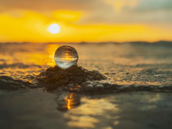 A glass sphere perched on a rock at the shore, reflecting the sunset over the calm, golden-tinted sea.