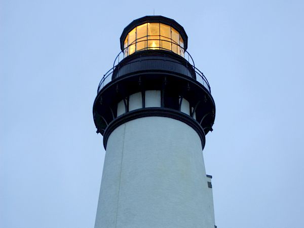 A tall white lighthouse with a glowing yellow lantern at the top against a pale blue sky.