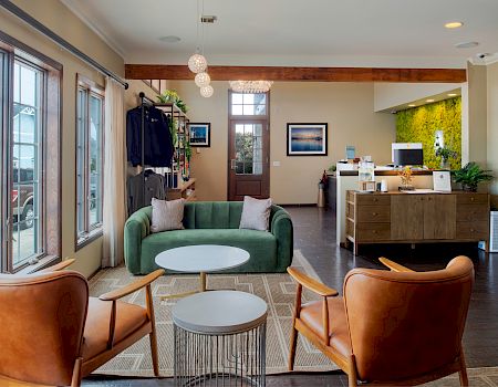 A stylish reception area with a green sofa, two orange leather chairs, round coffee tables, and a small white nesting table, plus a reception desk in the background.