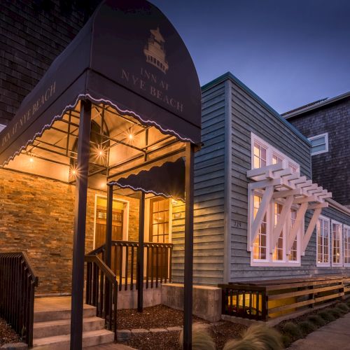 A cozy storefront at dusk with warm lights under a black awning, stone and wood exterior, a welcoming entrance, and a quiet street.