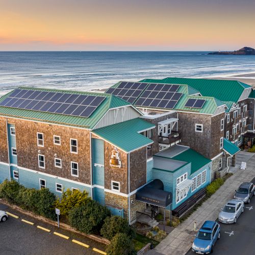 A seaside building complex with green roofs and solar panels along a coastal road, overlooking the ocean at sunset.