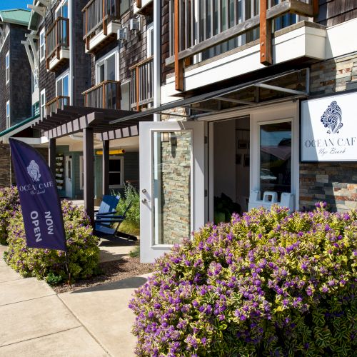 A storefront with stone facade, flower bushes, and a sign reading &ldquo;Chelsea&rdquo; or similar; a sidewalk leads to a glass door&mdash;sunny day.