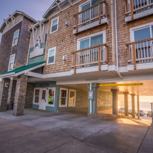 A sunny coastal motel with wooden balconies, stone columns, and a sheltered entrance; the warm sunset glows over the pier area.