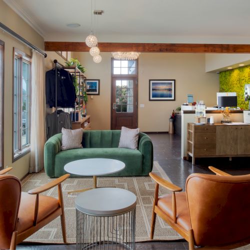 A modern, cozy lobby or lounge with a green sofa, orange chairs, round coffee tables, large windows, and a reception desk in the background.