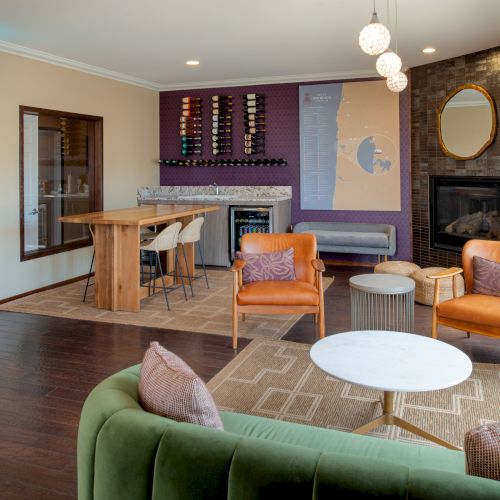 A modern, cozy living area with a green sofa, orange chairs, round coffee tables, a stone fireplace, and a small bar/desk area near large windows.