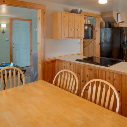 A sunny, cozy kitchen and dining area with wooden cabinets, a central island, a large wooden table and chairs, and pastel walls near a blue door.