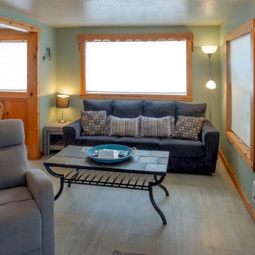 Cozy living room with a dark sofa, patterned throw pillows, a glass coffee table, a light gray recliner, and warm wood-trimmed windows and doorway, sunny and inviting.