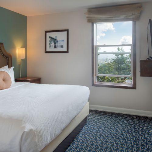 A bright hotel room with a large bed, nightstands, wall art, a window with sheer curtains, and a small desk near the light-filled corner.