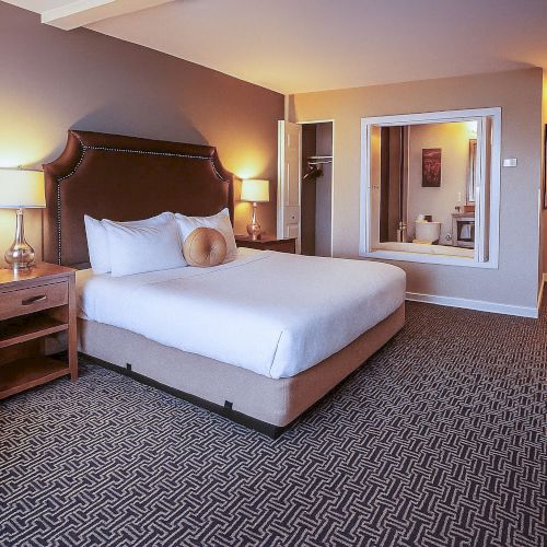 A cozy hotel room with a large bed, armchair, wooden furniture, warm lighting, and a small desk area.