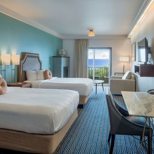 A modern hotel room with a large bed, blue decor accents, a desk, a seating area, and a bright, airy layout.