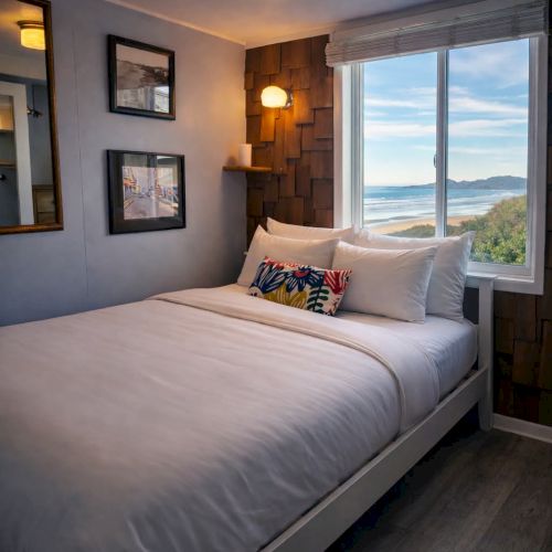 A cozy bedroom with a queen bed, soft gray bedding, framed art on the walls, warm lighting, and a large window overlooking water.