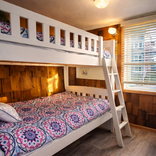 A cozy bunk bedroom with a white wooden bunk bed, colorful mandala bedding, a ladder, wood paneled walls, and a window with blinds. End.