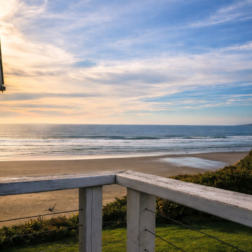 A serene beach at sunset with pastel skies, calm waves, and a railing in the foreground, inviting a peaceful coastal moment.