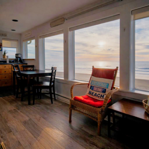 A cozy beachfront living space with large windows, a wood floor, a wicker chair with a beach-themed cushion, a small table, and a kitchen nook, overlooking the ocean.