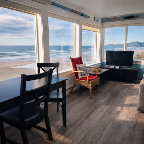 A bright coastal living area with large windows, ocean view, black chairs, a wicker chair with a red cushion, and a small TV stand.