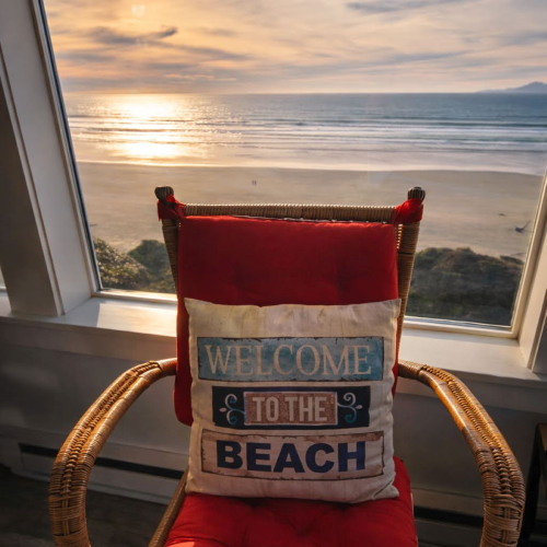 A red cushioned wicker chair by a large window overlooking a sunny beach at sunset, with a decorative &ldquo;Welcome to the Beach&rdquo; pillow.