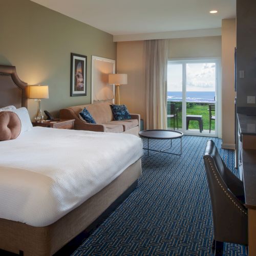 A cozy hotel room with a large bed, desk, and seating area; warm lighting, a flat-screen TV, and a window with daylight streaming in.