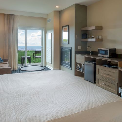 A cozy, modern apartment living area with a bed on the left, a kitchen to the right, and a bright, airy feel with natural light from large windows.
