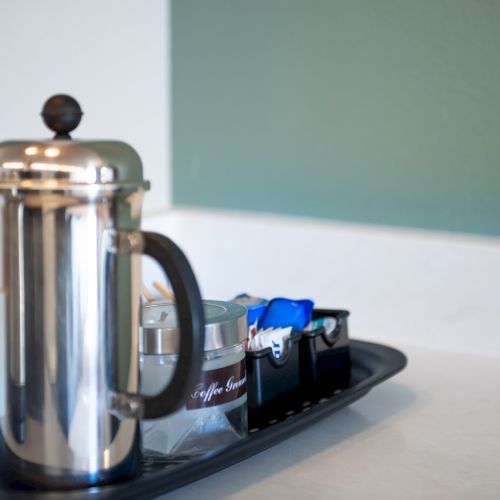 A polished coffee press and a tray with sugar packets on a light kitchen counter, ready for a morning brew.