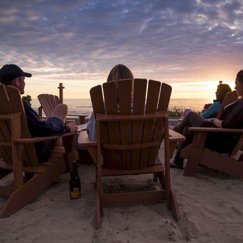 A group of friends sits in wooden beach chairs around a sunset on the sand, relaxing with drinks and conversation by the shore.