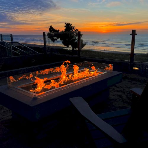 A coastal sunset with a fiery rectangular fire pit, glowing orange flames, sand, and a wooden deck railing overlooking the calm sea.
