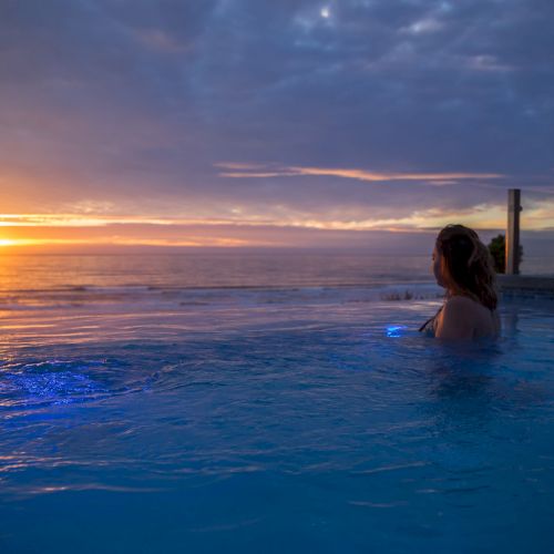 A person relaxing in a swimming pool at sunset, with calm water, a colorful sky, and a few posts or pillars along the pool edge, ending with a horizon glow.