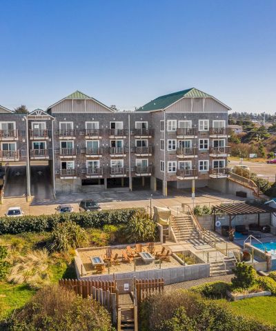 A row of three-story beach houses with balconies, a fenced yard, and a pool under construction in the foreground, set on a sunny coastal lot.