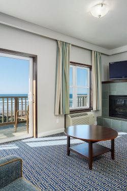 A bright living room with a view of the ocean, two chairs, a sofa, a coffee table, a fireplace, and sliding doors to a balcony.