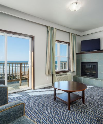 A bright living room with a view of the ocean, two chairs, a sofa, a coffee table, a fireplace, and sliding doors to a balcony.