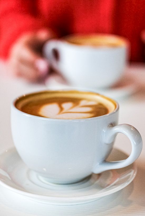 Two white cups of coffee with latte art on a white table; a person in a red top holds the back cup in the background.