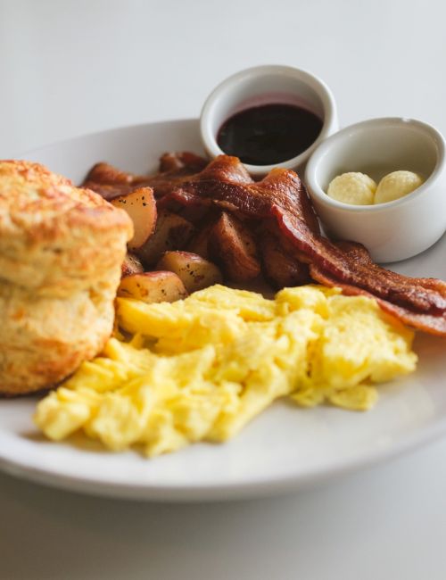 A breakfast plate with biscuits, scrambled eggs, crispy bacon, potatoes, sausages, and small cups of gravy and syrup, served on a white plate.