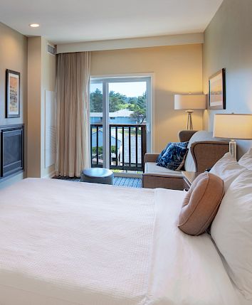 Cozy hotel room with a king bed, work desk, kitchenette, TV, fireplace, and balcony overlooking trees, bright and modern.