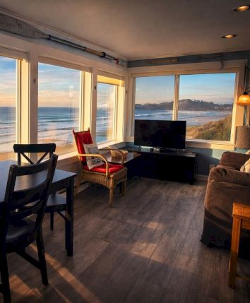 Cozy coastal living room with large windows framing a sunset over the ocean, a dining table, chairs, and a seating area facing the view.