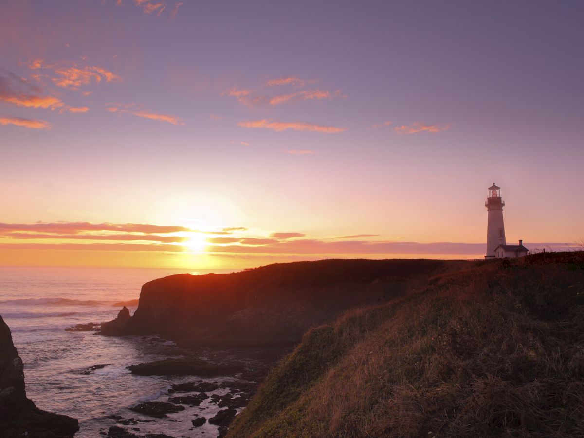 A sunlit coastline at sunset with a lighthouse perched on a cliff, the sea waves glinting as the horizon glows brilliantly.