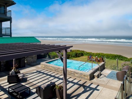 A sunny beach house with a pool, deck, and ocean view; people relax near the water as waves roll in.