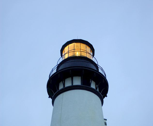 A tall white lighthouse with a glowing lantern room atop, guiding ships at dusk, its beacon spreading warm light over calm seas.