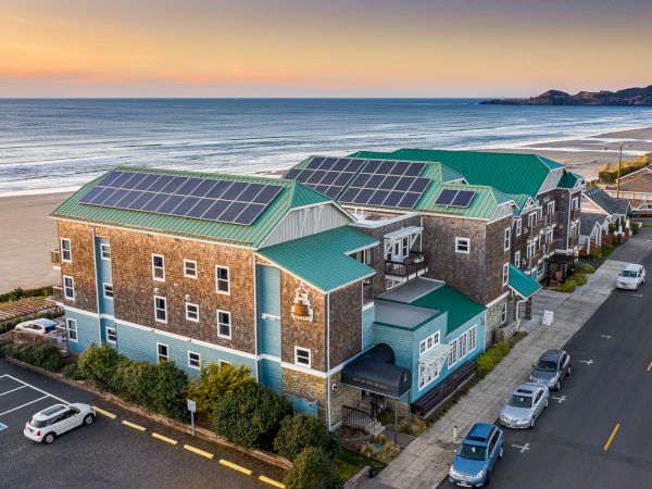 An aerial view of a seaside motel or hotel with solar panels on green roofs, brick walls, ocean backdrop, and a nearby street with parked cars.