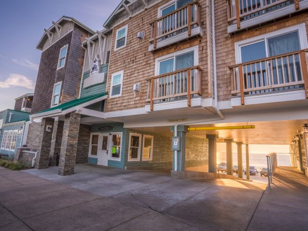 A beachfront hotel with brick and wood siding, balconies, and a covered entrance; sun sets over a boardwalk-like walkway at right.