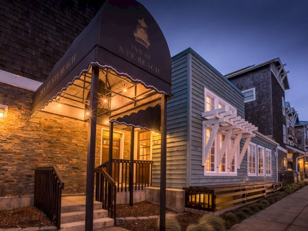 A warmly lit street of shops with stone and wood facades, a welcoming entrance canopy, and a quiet evening glow along the sidewalk.