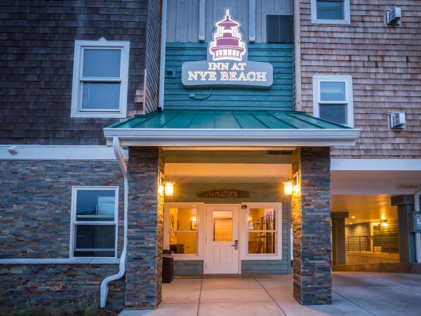 A hotel-style building entrance with a sign reading &ldquo;Inn at NYB Beach&rdquo; above a covered doorway, warm lights, stone and wood exterior, and a sidewalk.